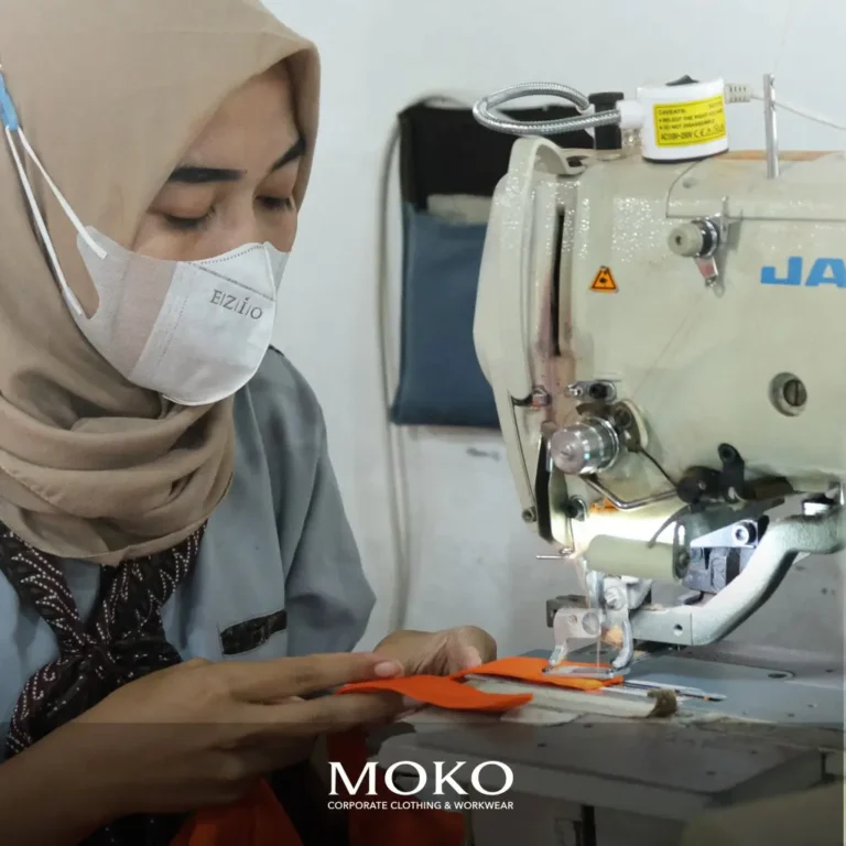 Seorang penjahit wanita dari Moko Konveksi sedang fokus menjahit seragam kerja berwarna oranye dengan mesin jahit industri.